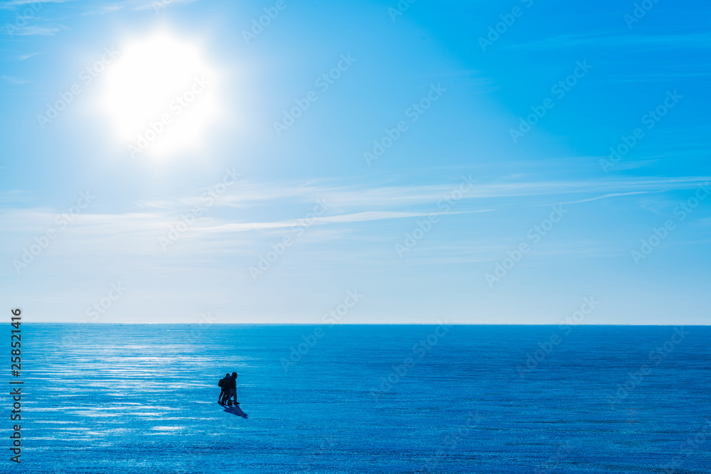 People coming on ice across the river or the lake in the winter sunny day. Blue winter landscape with bright Sun and clear sky with clouds