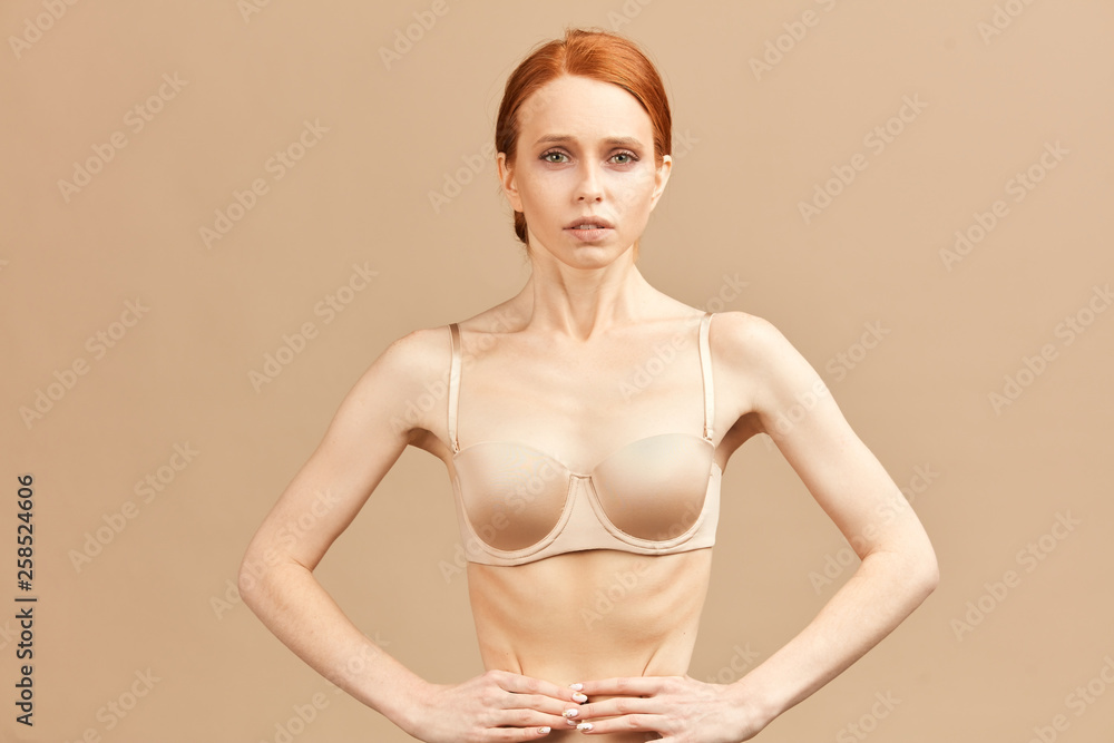 Foto de Skinny suffering female in nude underwear tied her waist with  measuring tape posing over beige background in studio. Anorexia and eating  disorders do Stock
