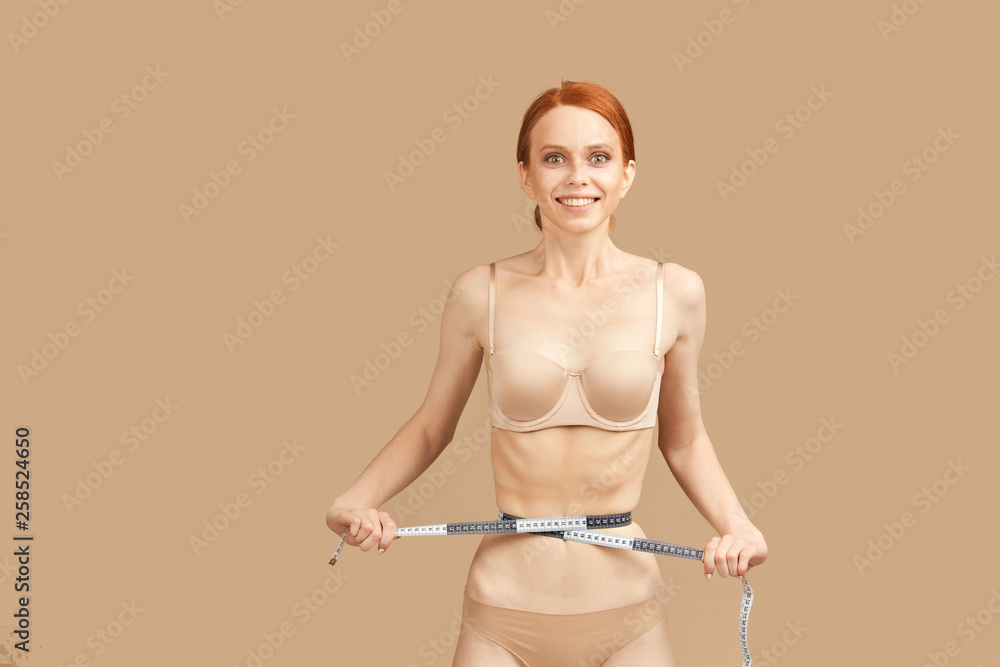 Woman suffering eating disorders anorexia or bulimia in nude bra and panties celebrates diet results, measuring waist with tape over beige background Woman suffering eating disorders anorexia or bulimia in nude bra and panties celebrates diet results, measuring waist with tape over beige background