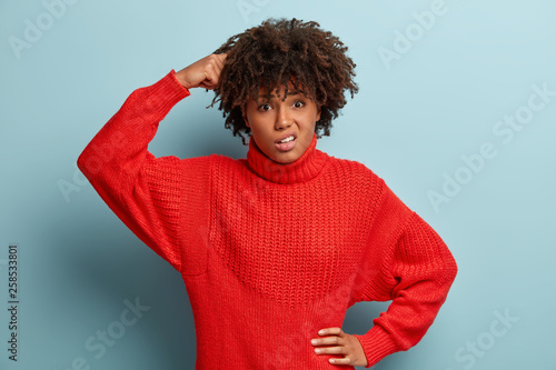 Confused thoughtful hesitant curly woman scratches head, keeps one hand on waist, looks with frustrated facial expression, has doubts, dressed in red knitted jumper, poses over blue background