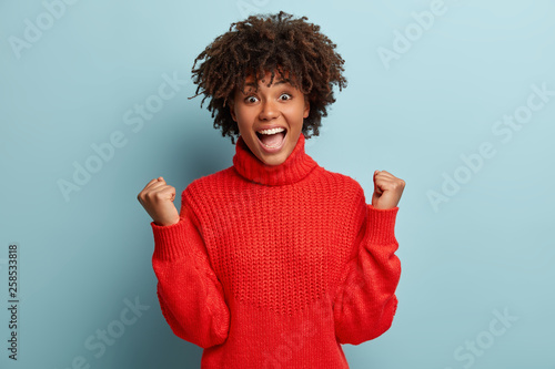 Glad young woman shows gesture of victory, wins competition, keeps fists raised, achieves desired result, screams from joy, wears red jumper, models over blue background. Reaching goal or aim