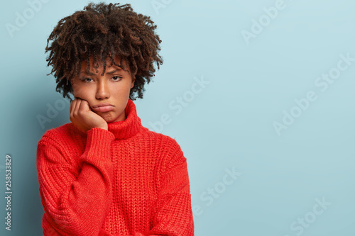 Human emotions concept. Displeased dark skinned woman with Afro haircut  gloomy expression  holds chin  feels sad and offended after quarrel with friends  wears red jumper  poses indoor alone