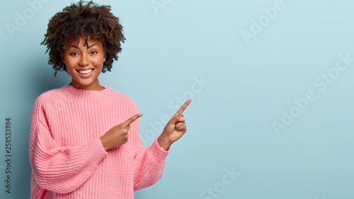 Quadro su tela Photo of cheerful young Afro American woman with satisfied expression, points aw