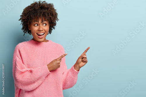 Foto Horizontal shot of positive lovely woman with Afro haircut, points away with both fore fingers, demonstrates copy space for your information, has appealing look, sincere smile on face