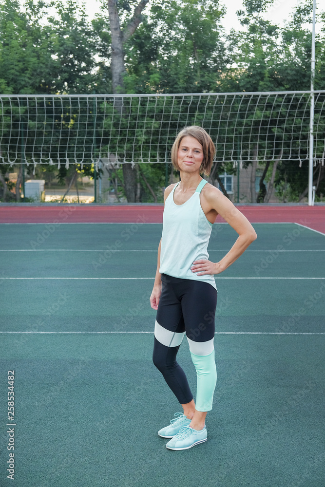 A woman in sports clothes and a yoga mat looks straight at the camera. A woman after a morning workout on the playground. A woman holding a gym mat and practicing yoga.
