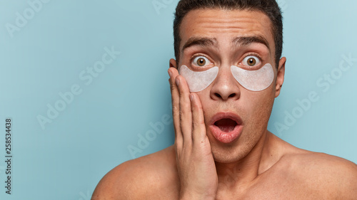 Personal care at home. Young stupefied man of European appearance keeps mouth widely opened, touches cheek, has patches under eyes, scared of something, poses bare shoulders over blue background