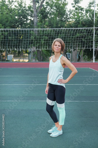 A woman in sports clothes and a yoga mat looks straight at the camera. A woman after a morning workout on the playground. A woman holding a gym mat and practicing yoga.