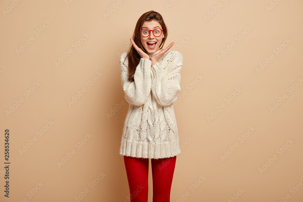 Glad lovely woman has happy expression, keeps hands near face, laughs at funny story, gazes at camera with joy, wears white long sleeved sweater and red leggings, isolated over beige background Glad lovely woman has happy expression, keeps hands near face, laughs at funny story, gazes at camera with joy, wears white long sleeved sweater and red leggings, isolated over beige background