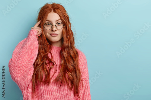 Photo of attractive ginger pupil with long wavy hair, tries to memorize something, keeps fore finger on temple, reminds information for exam, wears spectacles and jumper, stands over blue wall photo
