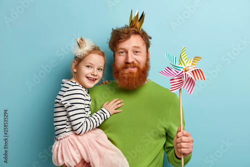 Happy ginger family of father and daughter wear party accessories, hold striped toy windmill, celebrate Fathers day or birthday, isolated over blue background. Fatherhood and celebration concept photo