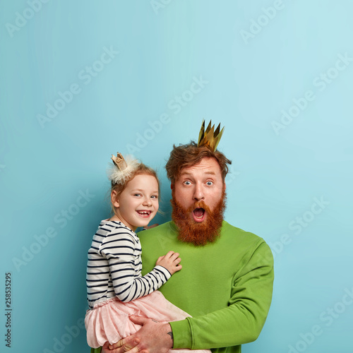 Canvas Print Vertical shot of surprised bearded red haired father carries small daughter, celebrates childs birthday, shocked to see many guests, have family celebrations