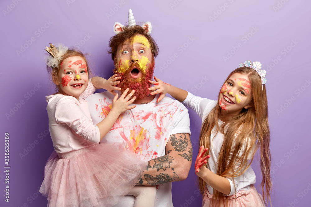 Φωτογραφία Funny two girls paint fathers beard with colorful paints, have fun together, celebrate holiday, wear festive apparel, have ginger hair, look alike, stand over purple background