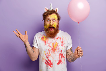 Emotional male has image of unicorn, gestures actively, has surprised expression, dirty face and t shirt, exclaims emotionally, holds balloon, entertains guests on party, suggests creative contest