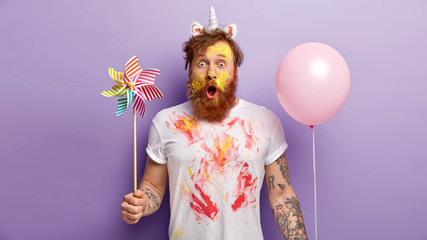 Indoor shot of surprised red man stares with shock, holds balloon and windmill in both hands, wears unicorn headband with horn and ears, smearked with colorful paints, entertains children on party