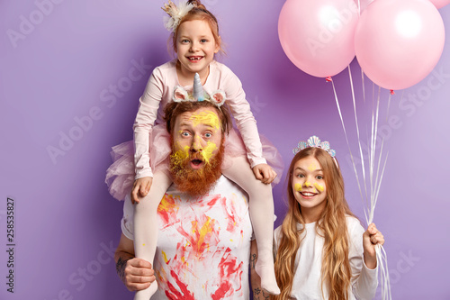 Surprised man entertains kids and organizes real holiday on Children day, being dirty with oil paints, piggybacks his little child lovely girl with crown holds air balloons. Party time and fun concept