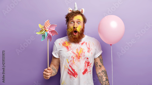 Indoor shot of surprised red man stares with shock, holds balloon and windmill in both hands, wears unicorn headband with horn and ears, smearked with colorful paints, entertains children on party
