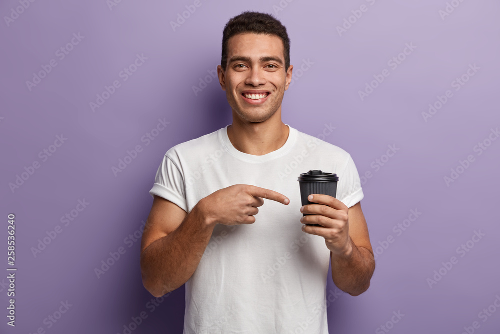 Youngster with toothy smile, wears casual white t shirt, points with fore finger at takeaway coffee, suggests to have drink, has pleasant appearance, isolated over purple background. Time for drink Youngster with toothy smile, wears casual white t shirt, points with fore finger at takeaway coffee, suggests to have drink, has pleasant appearance, isolated over purple background. Time for drink