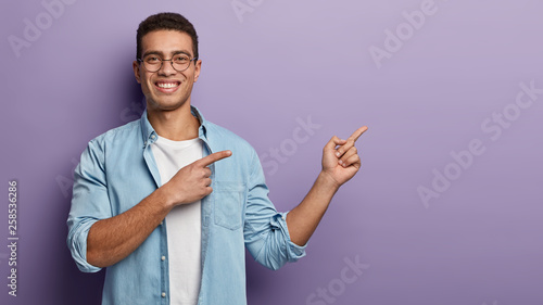 Positive glad handsome guy points rightside on blank copy space, smiles happily, dressed in blue shirt, promots object, isolated over purple background, shows direction, indicates at upper corner photo
