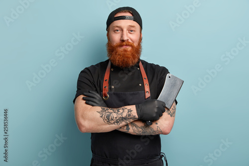 Portrait of handsome professional male butcher has arms crossed, holds metal sharp cleaver, wear black uniform, ready to start work, isolated over blue background. People and occupation concept photo