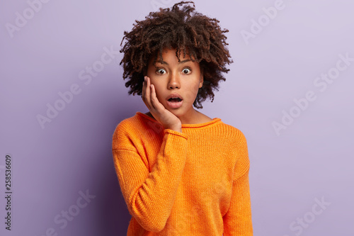 Facial expressions concept. Stupefied attractive woman keeps jaw dropped  touches cheek  hears astonishing news  can not believe rumors  wears casual orange knitted jumper  isolated over purple wall