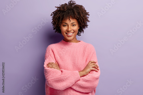Photo of self confident pleased dark skinned woman keeps arms folded, smiles gladfully while watches funny programme, has natural appealing beauty, wears oversized pink jumper, poses indoor. photo