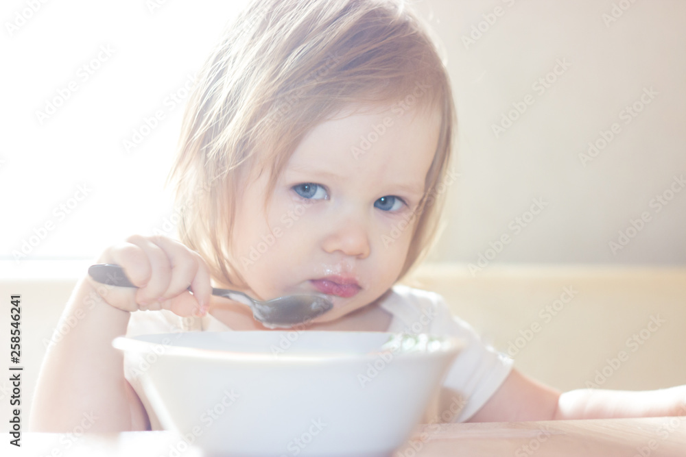 Little shaggy baby girl with pleasure alone holds a spoon and eats delicious food from a white plate. Little shaggy baby girl with pleasure alone holds a spoon and eats delicious food from a white plate.