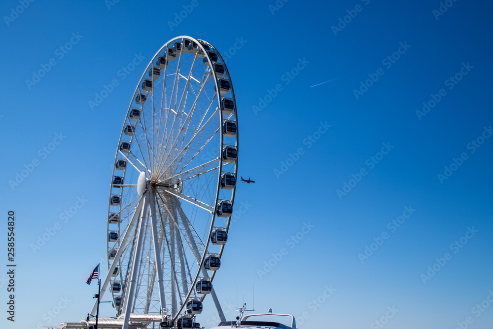 Airplane flies past a ferris wheel on a clear blue day, depicting vacation