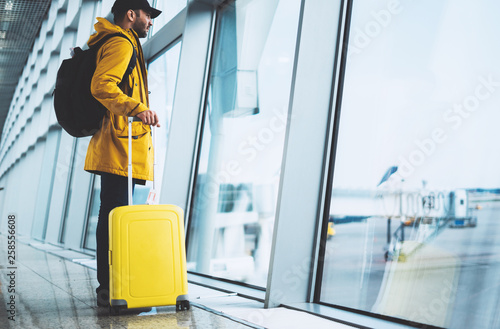 traveler with yellow suitcase backpack at airport on background window blue sky, passenger waiting flight in departure hall of lobby terminal lounge area, vacation trip concept, empty space mockup