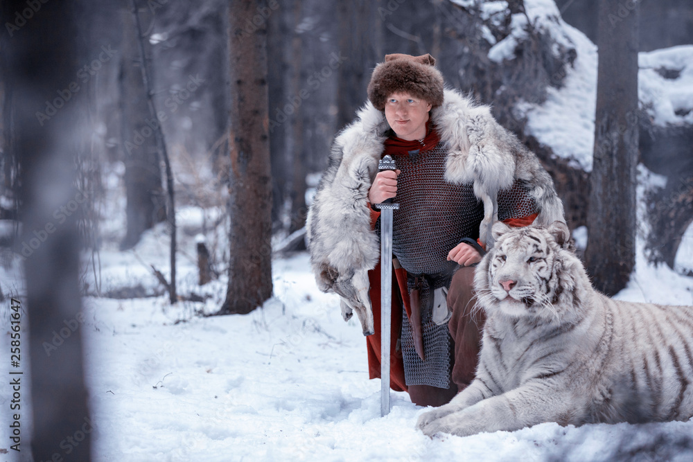 Man dressed as a medieval warrior stands in winter forest on one knee next to a white tiger holding a sword in his hand. The warrior is dressed in chain mail, a fur hat, a wolfskin and a red cloak.