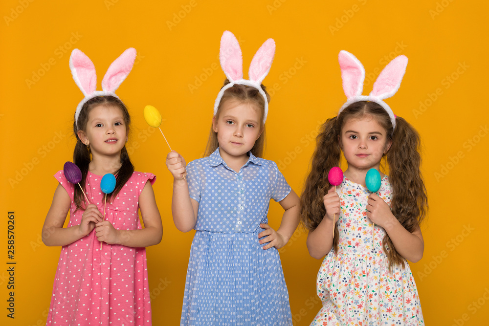 three little girls with Easter bunny ears holding colorful eggs on yellow background. Happy easter. cute children three little girls with Easter bunny ears holding colorful eggs on yellow background. Happy easter. cute children