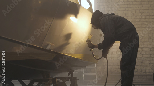 In slow motion a professional man (guy) in a garage (service) in working clothes dressed in a sweater sets up a paint gun (spray gun) and starts painting (putting a layer of ceramics) a yacht.