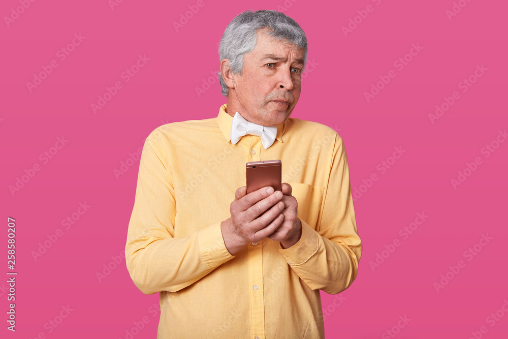 Φωτογραφία Close up portrait of mature man holding his smart phone in hands, does not know how to make video call, wears yellow shirt and white bowtie