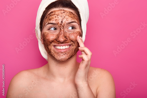Indoor shot of young woman applies face cosmetic mask, has toothy smile, has wrapped towel on head, poses half naked, looks at right upper corner, isolated over pink background. Skin care concept. photo