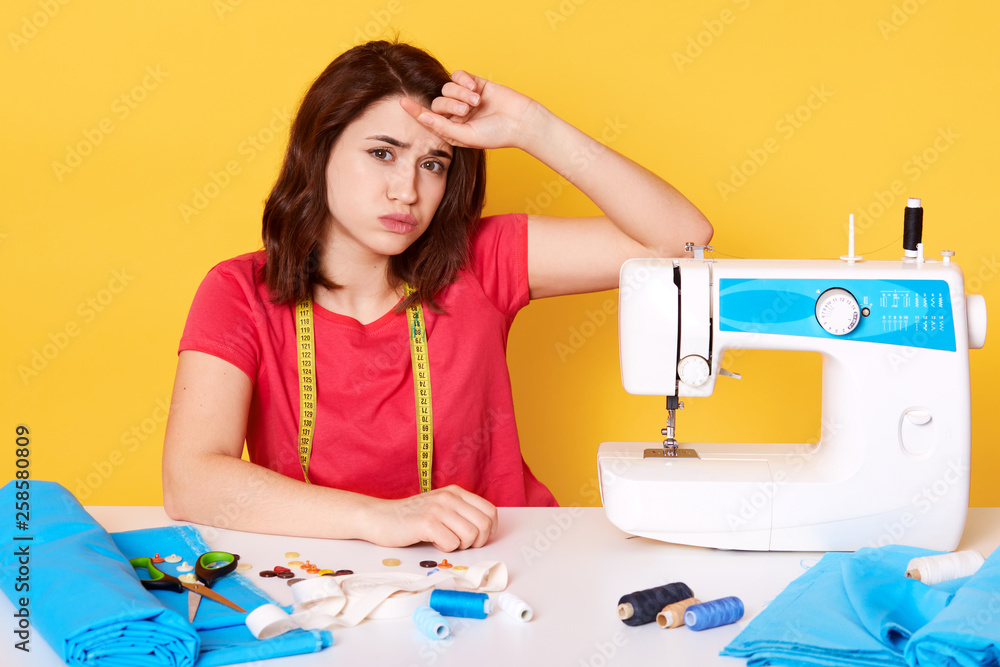 Image of good looking brunette female sits at working desk with sewing machine, nedles, thread, keeps hand on her forehead, being tired of sewing dresses, wears red t shirt, tapes measure on neck. Image of good looking brunette female sits at working desk with sewing machine, nedles, thread, keeps hand on her forehead, being tired of sewing dresses, wears red t shirt, tapes measure on neck.