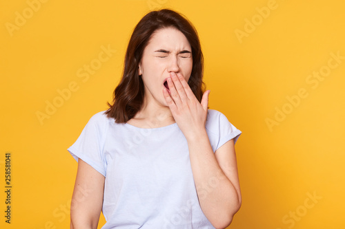 Beautiful woman yawns isolated over yellow background, wears casual white t shirt, has wavy hair, keeps hand near mouth. Free space for advertisment or promotional text. People and lifestyle concept.