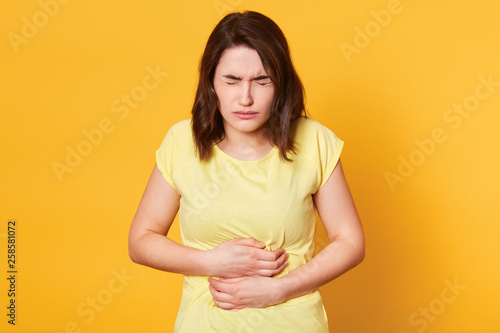 Close up portrait of young Caucasian woman with awful stomachache on yellow background, eats something expired, has intoxication, stands with closed eyes and keeps hamd on belly. Health concept. photo