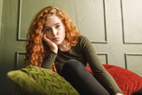 Curly redhead girl propped up her head and sitting at the sofa around the pillows in the green interior