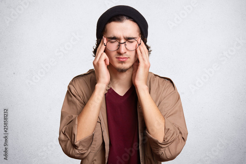 Studio shot of attractive unshaven male with closed, keeps fingers on temples, has terrible headache, wears stylish cap and shirt, isolated over white, background. People and lifestyle concept. © sementsova321