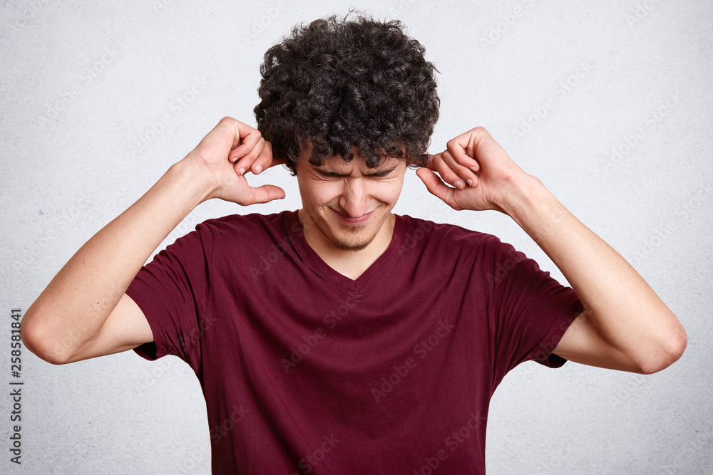 Portrait of worried handsome bearded man in casual maroon t shirt, standing with closed eyes and putting fingers in ears, does not want to hear anything, model poses isolated on white background. Portrait of worried handsome bearded man in casual maroon t shirt, standing with closed eyes and putting fingers in ears, does not want to hear anything, model poses isolated on white background.