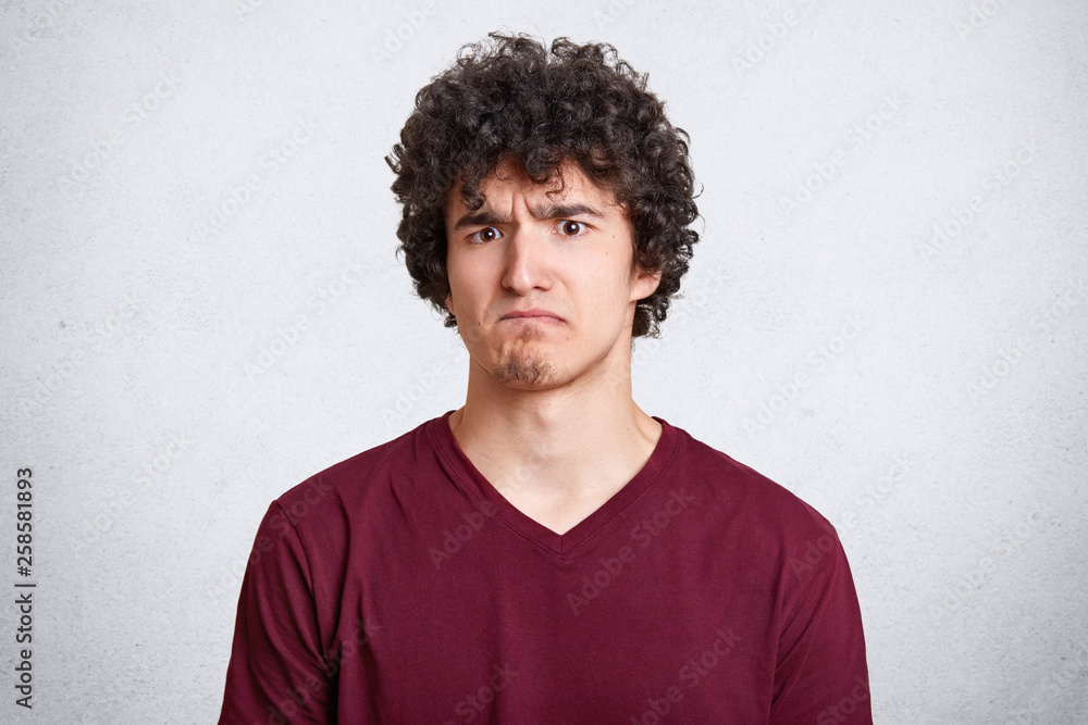 Gloomy bearded guy frowns face, being dissatisfied with results of exames, demands explanation why he looses. Handsome young man with dark curly hair isolated on white background. People and emotions Gloomy bearded guy frowns face, being dissatisfied with results of exames, demands explanation why he looses. Handsome young man with dark curly hair isolated on white background. People and emotions