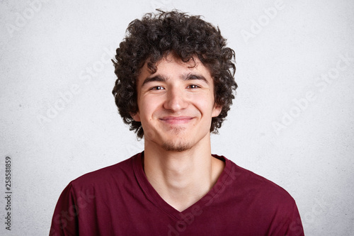 Photo of handsome unshaven guy looks with pleasent expression directly at camera, wears fashionable maroon t shirt, stands against white studio wall. People, lifestyle and fashion concept. Copy space. photo