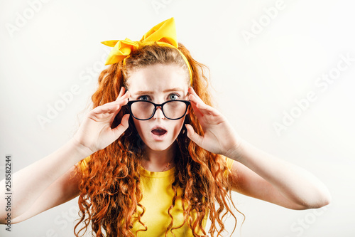 Surprised curly redhead girl in glasses with a yellow bow on her head wearing yellow t-shirt looking to the camera with schoked emotion