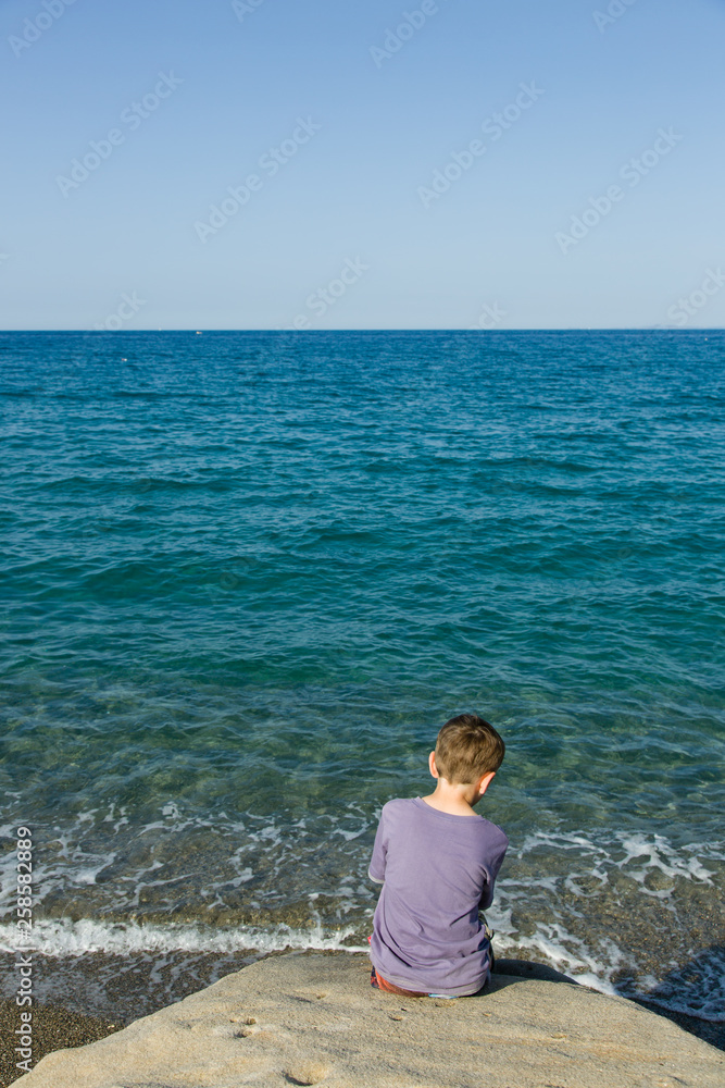 Blue sky and green - blue sea, horizon, silent waves and a little sitting boy can be seen from behind, in foreground coastal rock.