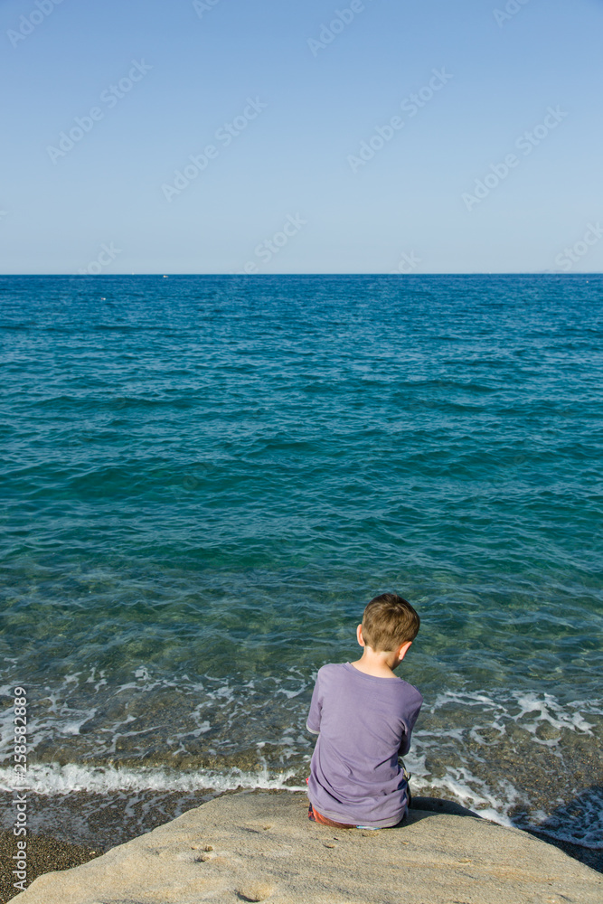Φωτογραφία Blue sky and green - blue sea, horizon, silent waves and a little sitting boy can be seen from behind, in foreground coastal rock