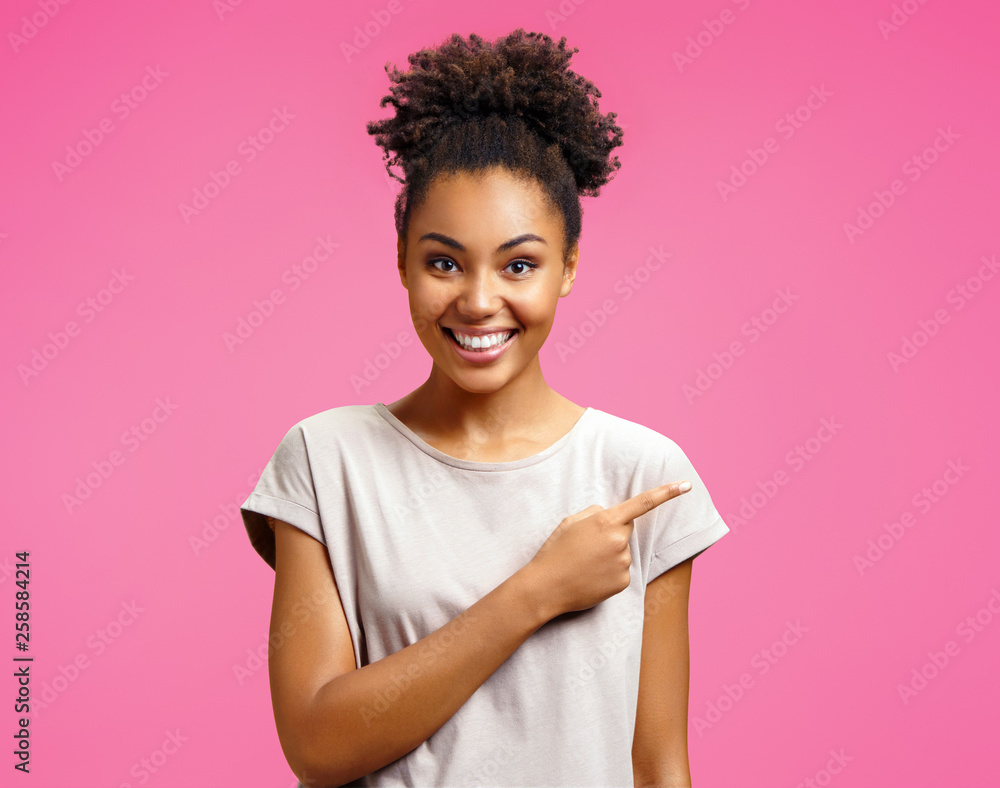 Overjoyed girl points aside, shows something at freespace. Photo of african american girl wears casual outfit on pink background. Emotions and pleasant feelings concept. Overjoyed girl points aside, shows something at freespace. Photo of african american girl wears casual outfit on pink background. Emotions and pleasant feelings concept.