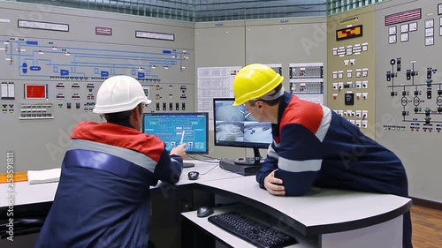 two engineers talk while looking at circuit and diagrams in monitors near main control panel of gas compressor station