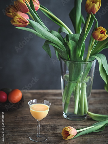 Colorful Easter eggs in nest, glass of egg nog and tulips in vase on wooden table.