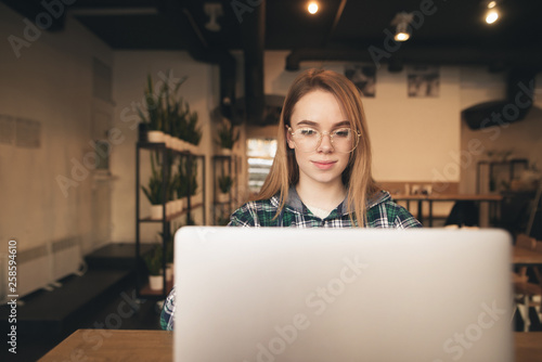 Attractive girl wearing glasses and a shirt uses a laptop in a cozy cafe, focuses on the screen and smiles. Teen girl with laptop sitting in a cafe.