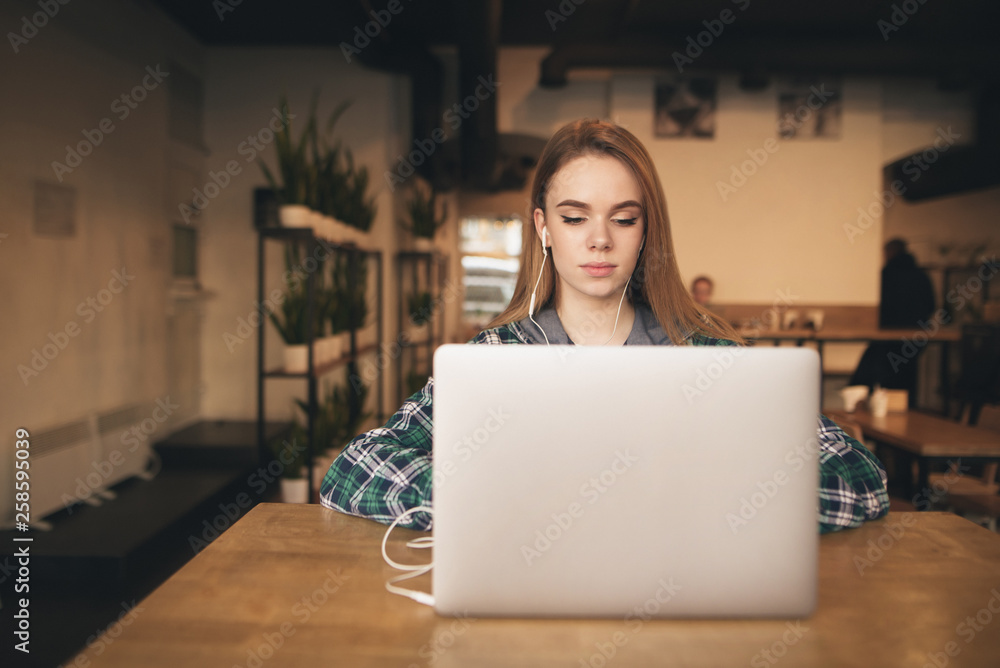 Beautiful student enjoys a laptop in a cozy coffee shop and is studying. Portrait of an attractive girl with a laptop in the cafe, listens to music in the headphones and looks at the screen. Beautiful student enjoys a laptop in a cozy coffee shop and is studying. Portrait of an attractive girl with a laptop in the cafe, listens to music in the headphones and looks at the screen.