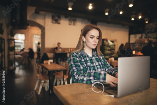 Cute girl works on a laptop in a cozy cafe and listens to music in headphones, wear casual clothing, she is focused on looking at the screen. Freelancer works in a cafe on a laptop.